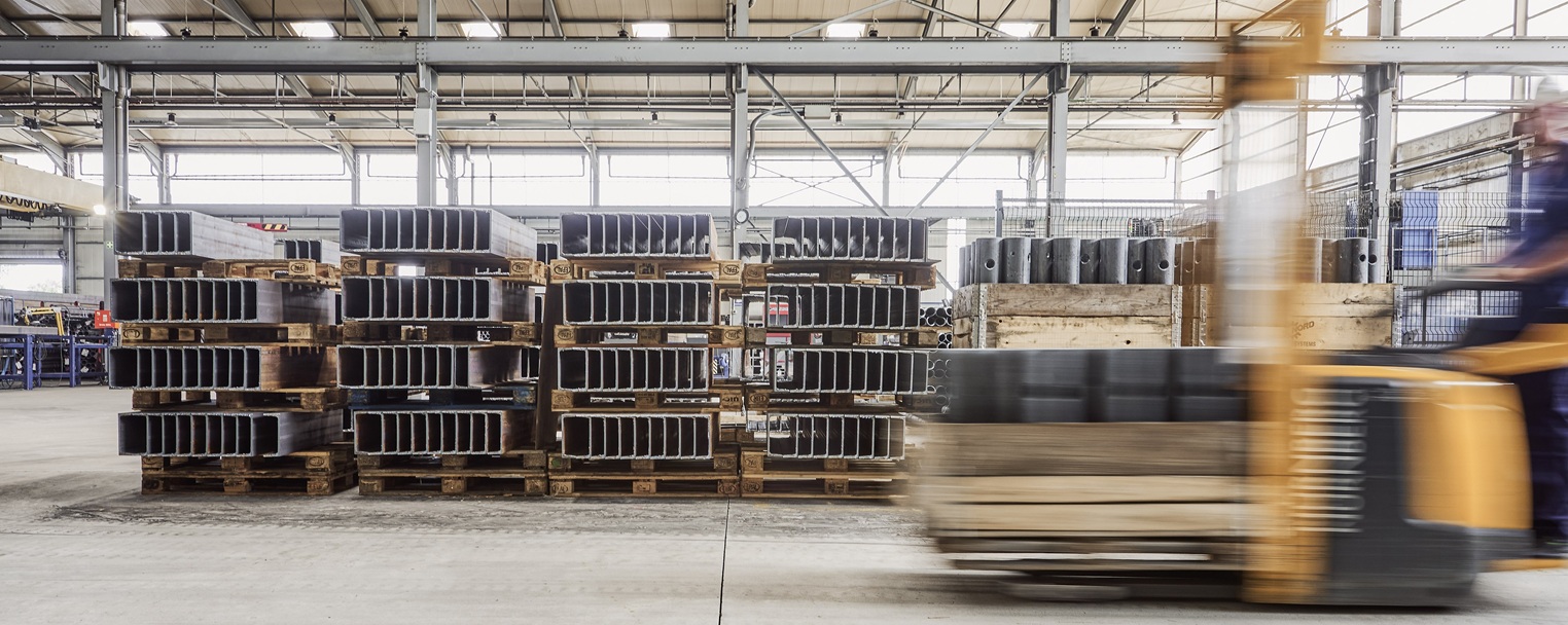 Gabelstapler fährt an gestapelten Metallträgern auf Holzpaletten in einem geräumigen Industrie-Lagerhaus mit hohen Decken vorbei.